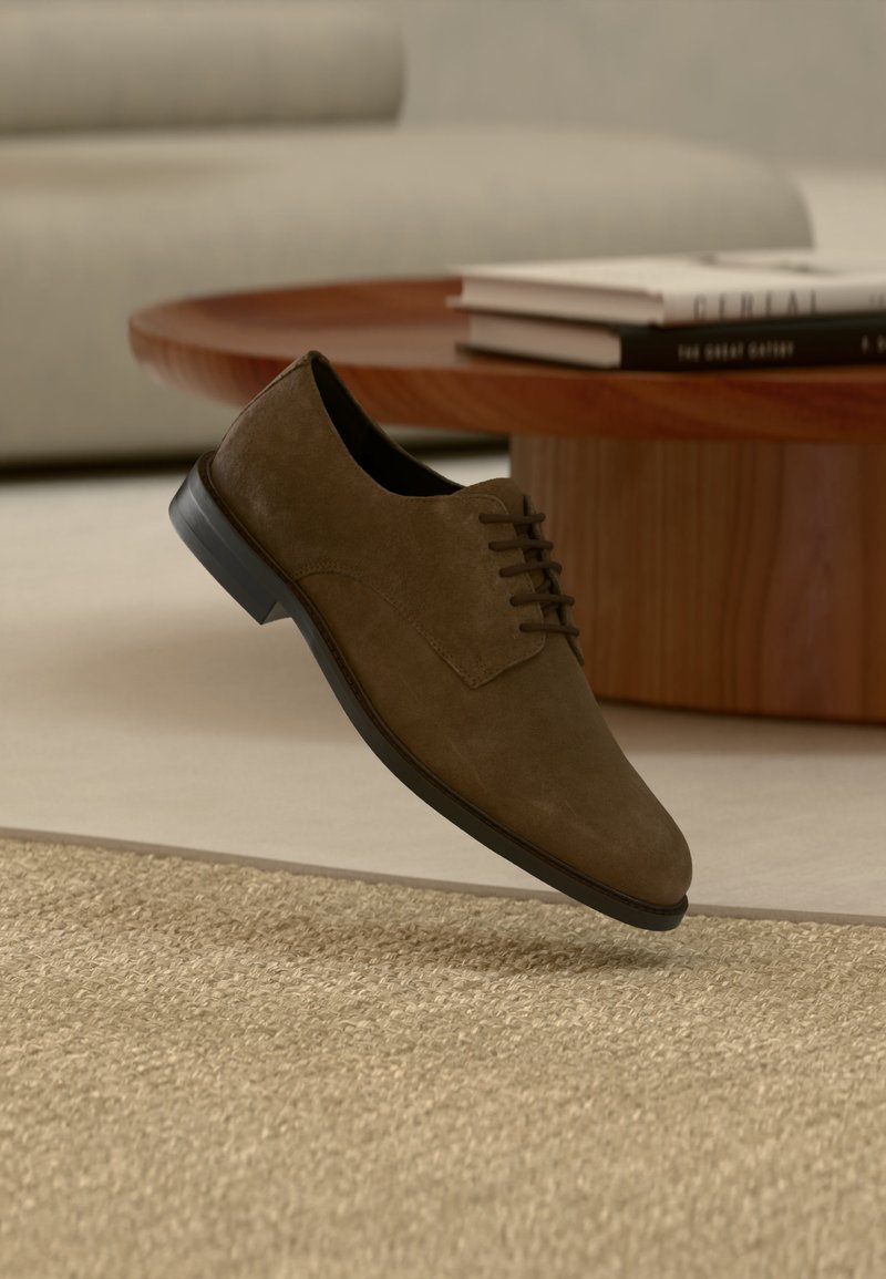Καφέ σουέντ ανδρικό επίσημο παπούτσι με μαύρη σόλα που αιωρείται πάνω από μπεζ υφαντό χαλί, δίπλα σε ξύλινο τραπεζάκι καφέ με στοίβες βιβλίων.