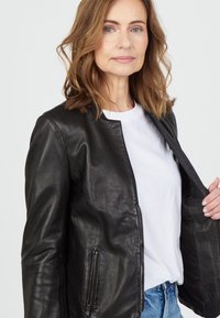 Zwarte leren jas, slim fit ontwerp, ronde kraag, zilveren ritsdetails aan de zijkanten, gedragen over een wit T-shirt. Soepele textuur, casual stijl.