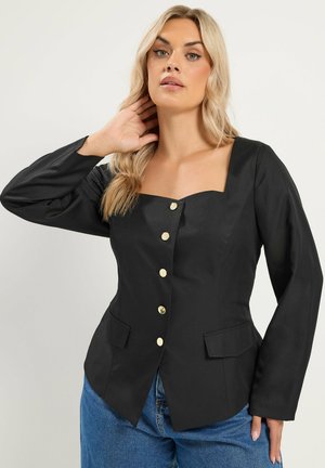 Femme aux longs cheveux blonds portant un blazer noir à col carré avec des boutons dorés et un jean bleu, posant la main près du cou.