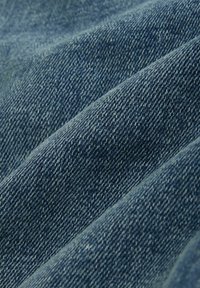 Tkanina dżinsowa w średnim odcieniu niebieskiego, z widocznym splotem o fakturze, widocznym przeszyciem i delikatnymi różnicami w kolorze.