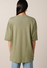 Olive zelené oversized tričko s krátkým rukávem. Měkká, hladká látka; kulatý výstřih; rovný lem. Kombinované s světle modrými džínami.