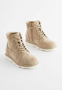 Beige suede nilkkurit nauhoilla ja sivuvetoketjulla, valkoisella kumisoletteella ja kontrastivärillä ommeltu yksityiskohta.