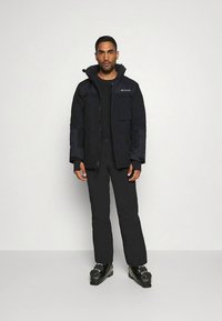 Marineblauwe waterdichte jas met ritssluiting, meerdere zakken en een capuchon, gedragen met zwarte broek en schoenen, staand tegen een witte achtergrond.