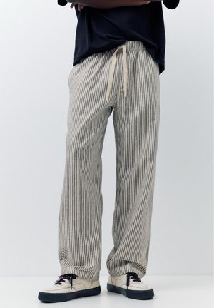 Osoba nosząca luźne beżowe i granatowe spodnie w pionowe paski z talią na sznurek oraz kremowe tenisówki z czarnymi podeszwami.