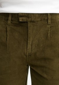 Pantalones de pana verde oliva con textura de estrías verticales, que presentan una cintura estándar y bolsillos frontales, aptos para un uso casual.