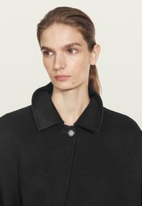 Ung person med lysebrunt hår trukket tilbage, iført en sort frakke med en stor krave, der kigger let mod venstre mod en ensartet lys baggrund.