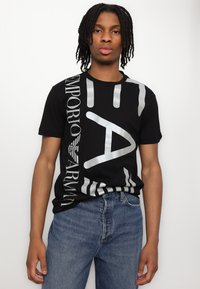 Tricou negru din bumbac, cu litere și grafice mari imprimate de culoare argintie. Asortat cu blugi din denim albastru. Fundal minim distragător.