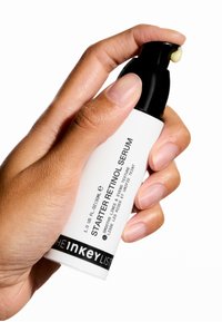 Main tenant un flacon pompe blanc de Sérum Rétinol Starter de The Inkey List avec de la crème distribuée sur la buse, sur fond blanc.