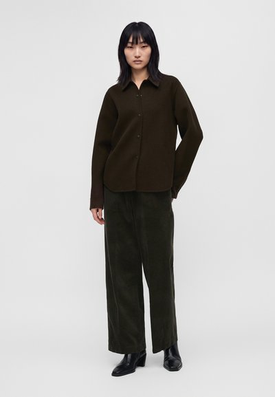 Femme aux cheveux noirs raides porte une chemise marron foncé boutonnée, un pantalon large foncé et des bottines noires à talons sur un fond clair uni.