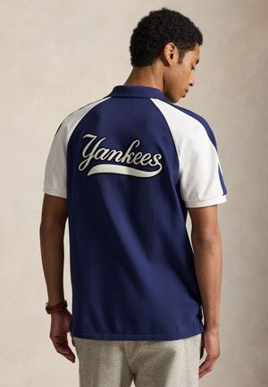 Egy férfi háttal áll, sötétkék és fehér galléros pólót visel, amelynek hátán a "Yankees" felirat hímzett, világosszürke nadrággal párosítva.