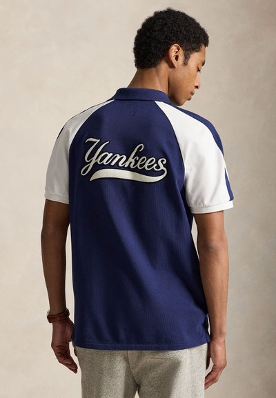Hombre de espaldas con una camisa polo azul marino y blanca con "Yankees" bordado en la espalda, combinado con pantalones gris claro.