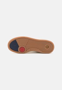 Semelle d'une basket avec des sections en caoutchouc beige, bleu marine et rouge, motifs antidérapants texturés, et « LACOSTE » embossé au centre.