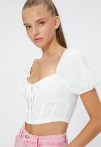 Top corto blanco con mangas abullonadas, tejido texturizado, escote cuadrado y detalle de lazo en el centro, combinado con pantalones cortos rosa claro.