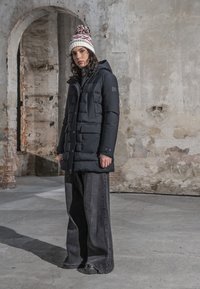 Femme portant une doudoune noire, un pantalon noir large et un bonnet tricoté avec un pompon, debout à l'intérieur contre un mur de briques usé.