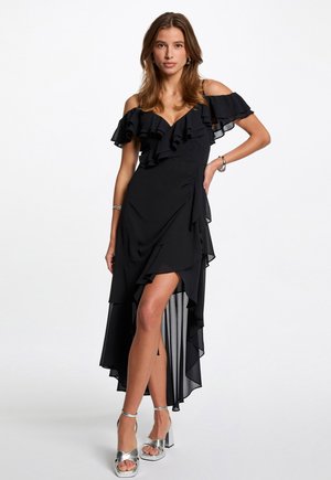 Donna che indossa un abito nero a spalle scoperte con volant e taglio asimmetrico, abbinato a sandali con tacco alto argentati, in piedi davanti a uno sfondo bianco semplice.