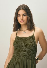Robe verte olive avec corsage froncé et jupe plissée. Elle présente des fines bretelles et une texture lisse et brillante. Collier en chaîne notable avec pendentif.