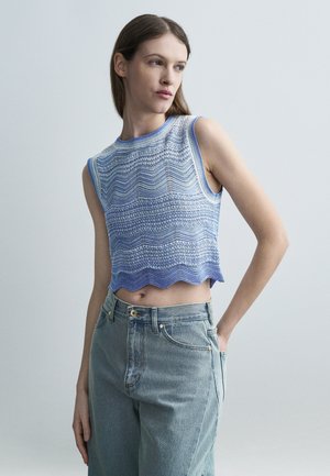 Vrouw die een mouwloze blauwe crop top met patroon draagt en lichtblauwe high-waisted jeans, staand met één hand in de zak tegen een effen achtergrond.