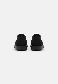 Schwarze Wildlederschuhe mit glatter Textur, abgerundeter Zehenpartie und durchbrochener Details im Obermaterial. Solide schwarze Gummisohle, minimalistisches Design.