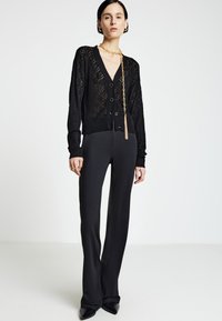 Cardigan nero in maglia con motivo a diamante, scollo a V e bottoni, abbinato a pantaloni neri svasati e scarpe nere a punta. Dettagli in catena dorata.