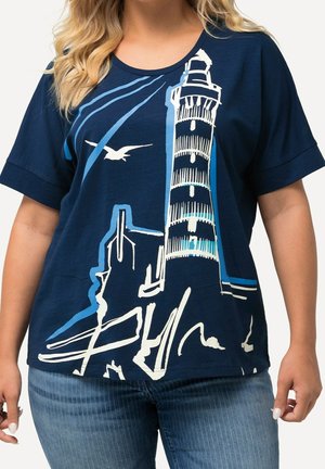 Kobieta w granatowym t-shircie z białą i jasnoniebieską ilustracją latarni morskiej i mewy, w połączeniu z niebieskimi dżinsami.