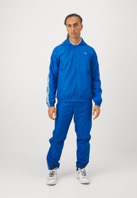 Kék tréningruha sima anyagból, zipzárral ellátott dzsekivel és rugalmas mandzsettákkal. Fehér részletek az ujjakon és a nadrágon, sportcipővel párosítva.