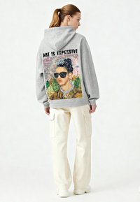 Felpa con cappuccio grigia oversize con una grafica colorata stampata sul retro con la scritta "L'ARTE È COSTOSA." Abbinata a pantaloni cargo color crema.