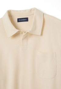 Beige Polohemd mit klassischem Kragen, Knopfleiste und einer kleinen Brusttasche. Hergestellt aus weichem Baumwollstoff mit strukturierter Oberfläche.