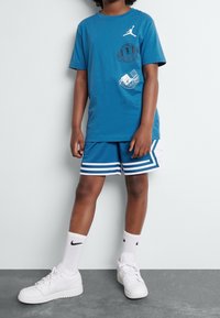 Modrá bavlněná tričko s logem Jordan, ve spojení s modrými síťovanými šortkami s bílými pruhy. Oblečeno s bílými teniskami Nike a krátkými ponožkami.