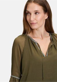 Blusa leggera verde oliva con scollatura a V, dettagli con rifiniture argento e maniche ampie con polsini elasticizzati. Tessuto con texture a righe verticali.