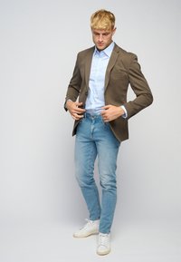 Brun blazer over en lysblå stribet skjorte, parret med lysblå jeans og hvide sneakers; tætsiddende design med glatte teksturer.