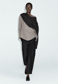Grijze linnen top met een asymmetrische zwarte gebreide omslag, gecombineerd met losse zwarte broeken en glanzende zwarte schoenen. Eenvoudige metalen hangersieraden.