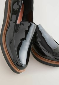 Melni lakotie kurpes ar spīdīgu virsmu, brūnu gumijas zoli un elegantu dizainu, kas ietver elastīgas sānu akcentus.