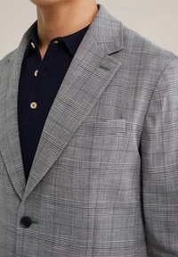 Lichtgrijze geruite blazer met een notch lapel, enkele knoopsluiting en textuurstof. Gedragen over een marineblauwe polo met knopen.