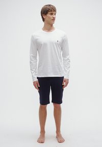 Hvit langermet skjorte med en liten logo på brystet, kombinert med marineblå shorts til knærne. Modellen står barfot mot en ensfarget bakgrunn.