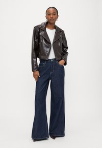 Brūna mākslīgās ādas saīsināta jaka ar sudraba rāvējslēdzējiem, kombinēta ar tumši zilām plašu kāju augsta vidukļa džinsa biksēm. Modele ir ģērbusi baltu topu zemāk.