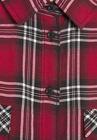 Chemise en flanelle à carreaux rouge et noir avec un col, comportant des boutons noirs et une poche poitrine avec un motif à carreaux. Tissu texturé.