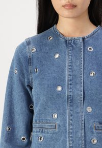 Denim jakna v svetlo modri barvi s srebrnimi zakovkami, sprednjim zapiranjem in dvema horizontalnima žepoma; gladka tekstura in klasična zasnova.