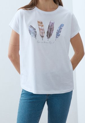 Personne portant un t-shirt blanc avec quatre plumes colorées et le texte "la vie est belle" imprimé sur la poitrine, associé à un jean bleu.