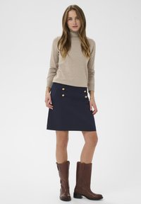 Pull à col roulé beige, jupe marine avec des boutons dorés, bottes en cuir marron montant jusqu'aux genoux, textures lisses, coupe ajustée.