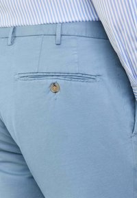 Lichtblauwe broek met een achterzak en knoopsluiting. De stof heeft een gladde textuur en een subtiel geweven patroon.