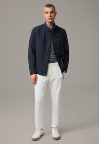 Tengerészkék steppelt bomber dzseki, álló gallérral, elülső cipzárral és zsebekkel, fehér slim-fit chino nadrággal és barna talpú fehér sportcipővel párosítva.