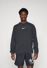 Sort Nike treningsgenser med lange ermer i svart med hvitt logo, elastisk midje; kombinert med svarte shorts med hvit prikkemønster og logo-detaljer.