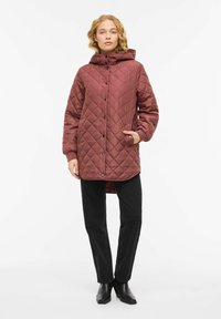 Quiltet burgunderrød jakke med hette, trykknapper og lommer. Kombinert med svarte bukser og ankelstøvler. Jevn tekstur og midtlengde design.