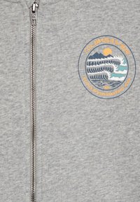 A hoodie cinza com fecho zip, apresenta uma graphic circular de onda do oceano em azul e verde, com um design de sol e montanha. Zíper de metal durável.