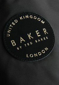 Sort cirkulær lædermærke med prægede guldtekst: "UNITED KINGDOM BAKER af TED BAKER LONDON" på en tekstureret sort baggrund.