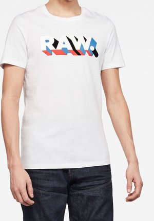 Weißes Kurzarmhemd mit dem Schriftzug "RAW." in großen, fetten Buchstaben in Weiß, Schwarz, Rot und Blau, getragen von einer stehenden Person in dunklen Jeans.