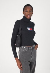 Maglione a collo alto nero a costine con toppe colorate. Abbinato a jeans grigi a vita alta e a una borsa a tracolla nera.