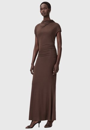 Kobieta o ciemnej karnacji z krótkimi włosami ubrana w dopasowaną, marszczoną, brązową suknię maxi z krótkim rękawem typu czapka oraz czarne sandały na wysokim obcasie z paskami, stojąca.