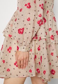 Robe beige texturée avec motifs floraux rouges et petits pois bruns, manches longues avec poignets et boutons décoratifs en perles, montrée sur une main reposant.