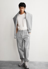 Grijze sweatpants met een tailleband met trekkoord en zijzakken, gecombineerd met een witte t-shirt met zwarte tekst. Lichtgrijze trui over de schouders gedrapeerd.